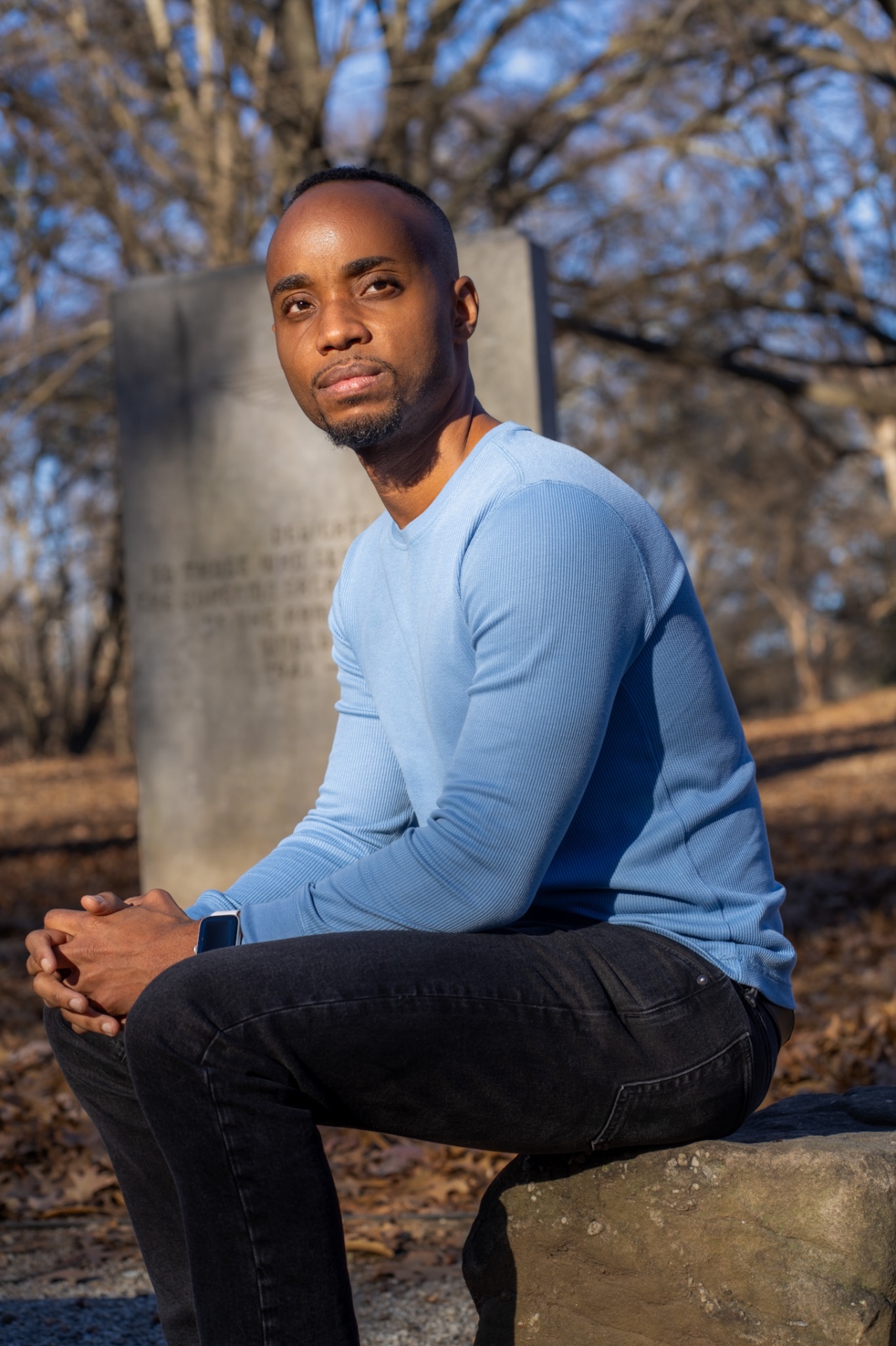 Outdoor portrait of a man in a light blue top seated on a stone in winter park light